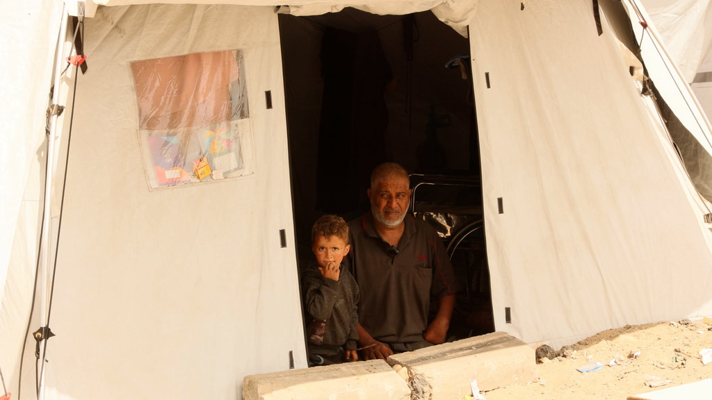 Un hombre y un niño pequeño están sentados en la entrada de una tienda de campaña blanca en un campamento de desplazados en Gaza.