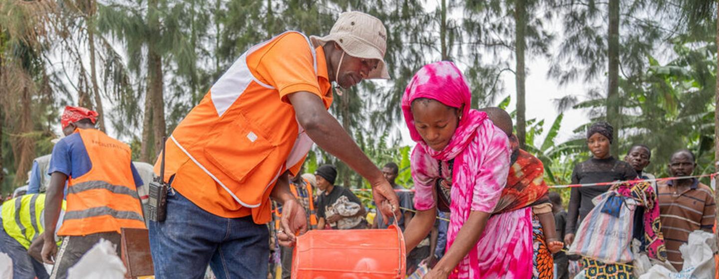 À Bweremana, en RDC, un employé du PAM vêtu d'un gilet orange verse des céréales d'un seau dans un sac blanc destiné à une femme portant un foulard rose, sur un site de distribution alimentaire.