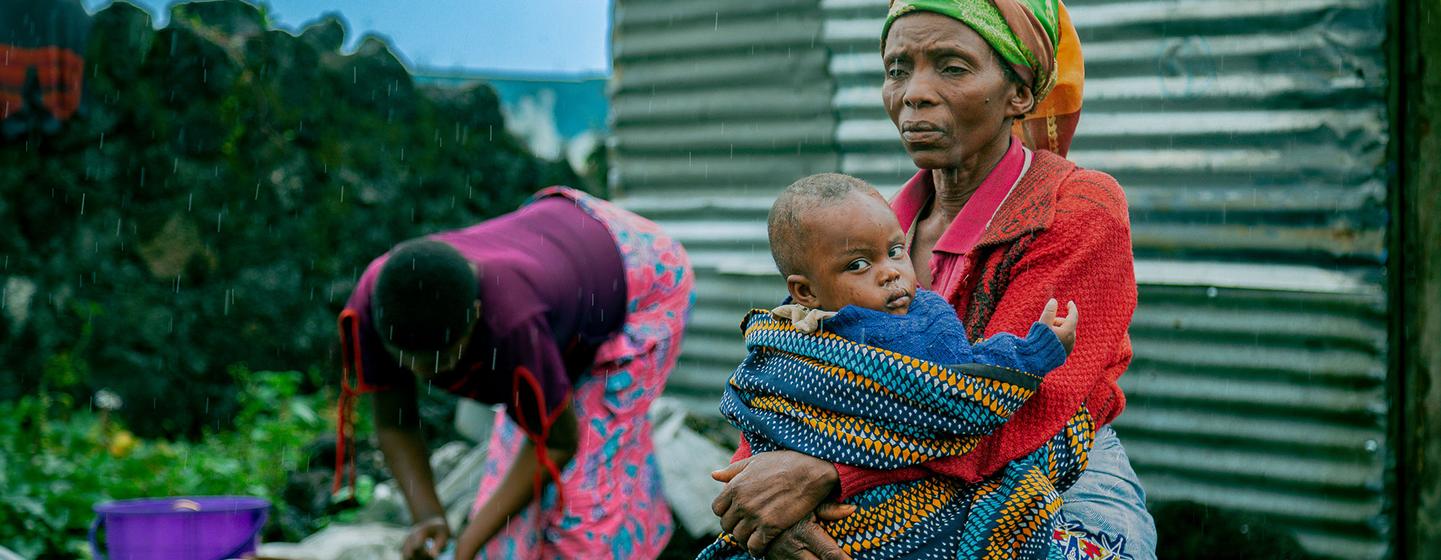 Une femme vêtue d'une veste rouge et d'un foulard est assise sur un banc de bois sous la pluie, tenant un bébé. En arrière-plan, une autre personne lave du linge dans une bassine.