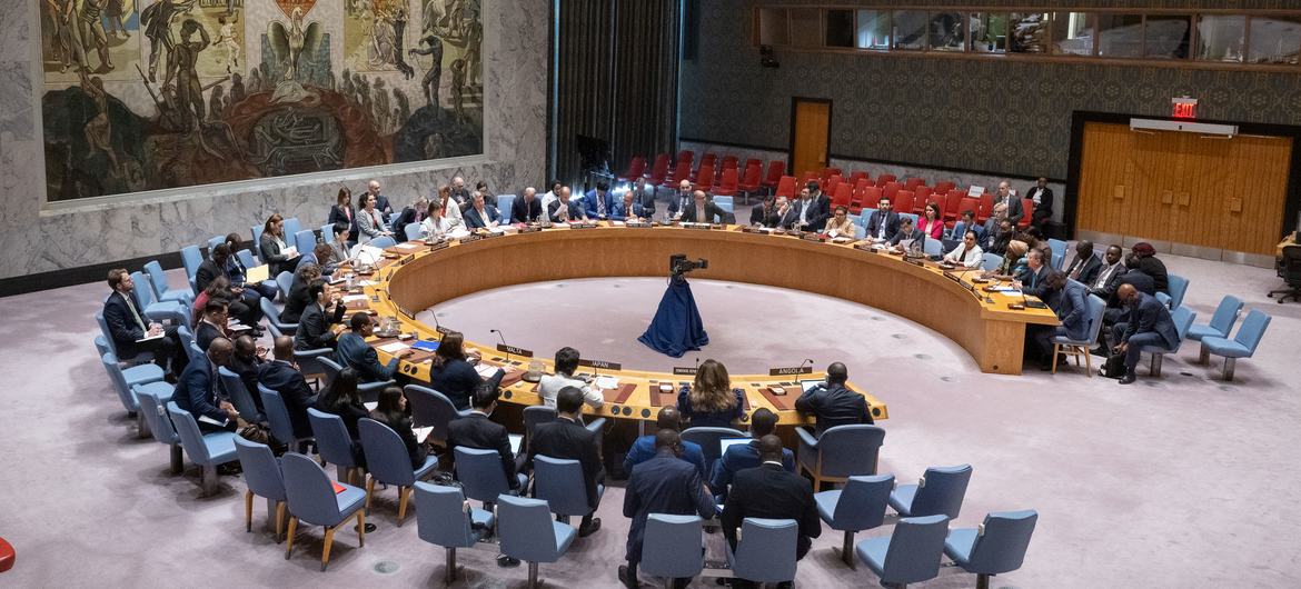 纽约联合国安理会会议的全景镜头，显示代表们围坐在圆桌旁，有人站起来作报告。
