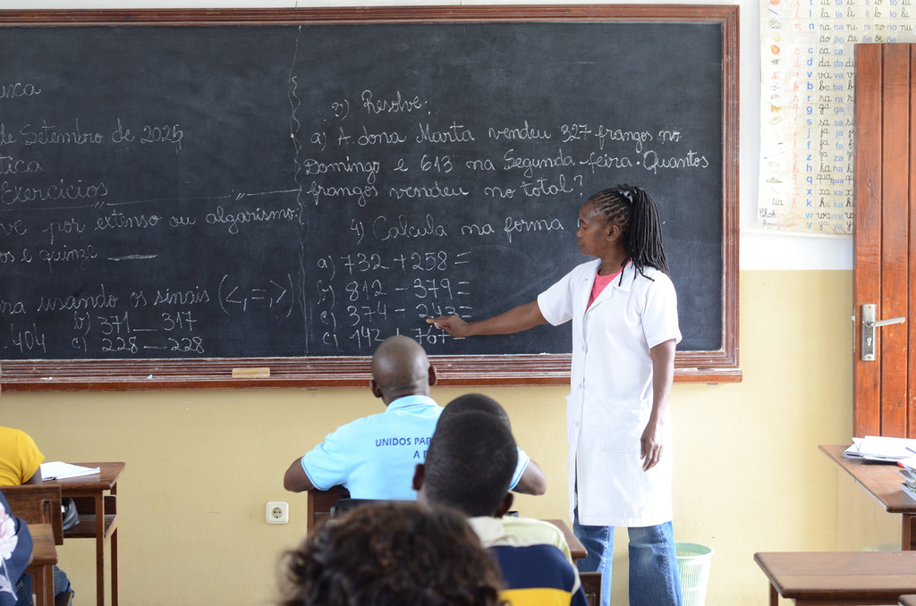 Uma professora de jaleco branco aponta para uma lição de matemática escrita em um quadro-negro em uma sala de aula em Moçambique, com alunos sentados em carteiras.