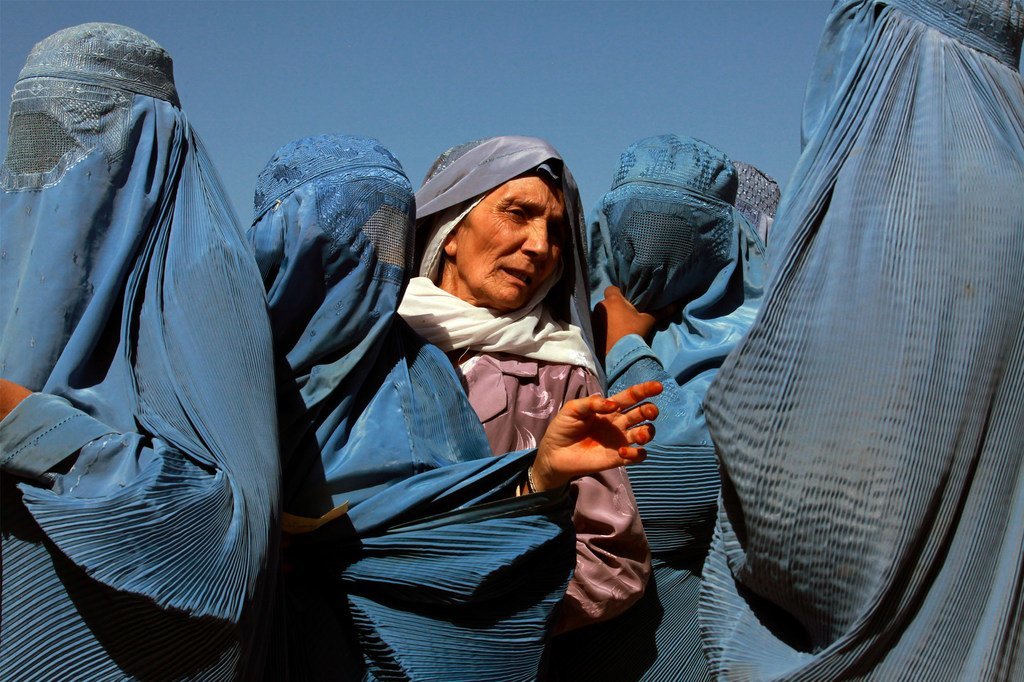 Uma idosa afegã com um lenço roxo na cabeça está rodeada por outras mulheres com burcas azuis num ponto de distribuição do Programa Mundial de Alimentos em Herat, Afeganistão.