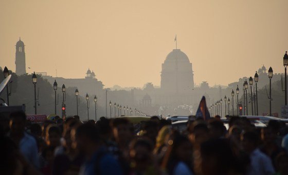A crowded street in New Delhi, India, is shrouded in thick smog, with the silhouette of a domed government building visible in the background.