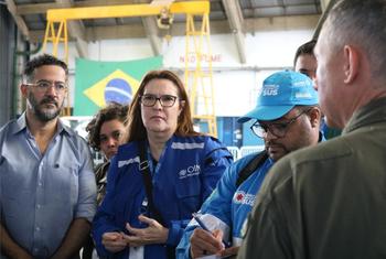A chefe de programas da OIM, Socorro Tabosa, posa com colegas durante uma reunião em uma instalação brasileira, com uma bandeira do Brasil ao fundo.