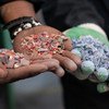 Mikono ikiwa imeshika vipande vya plastiki vyenye rangi na vipande vya plastiki vilivyosindikwa, huku taka za plastiki zikiwa nyuma.