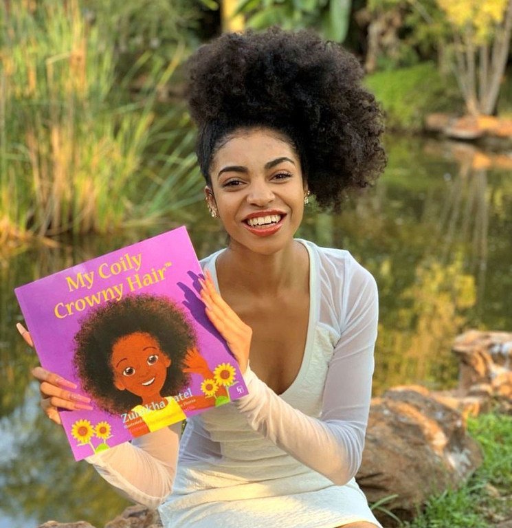 A ativista Zulaikha Patel exibe seu livro infantil, "My Coily Crowny Hair" (Meu Cabelo Encaracolado e Coroado), em um cenário ao ar livre perto de um lago.