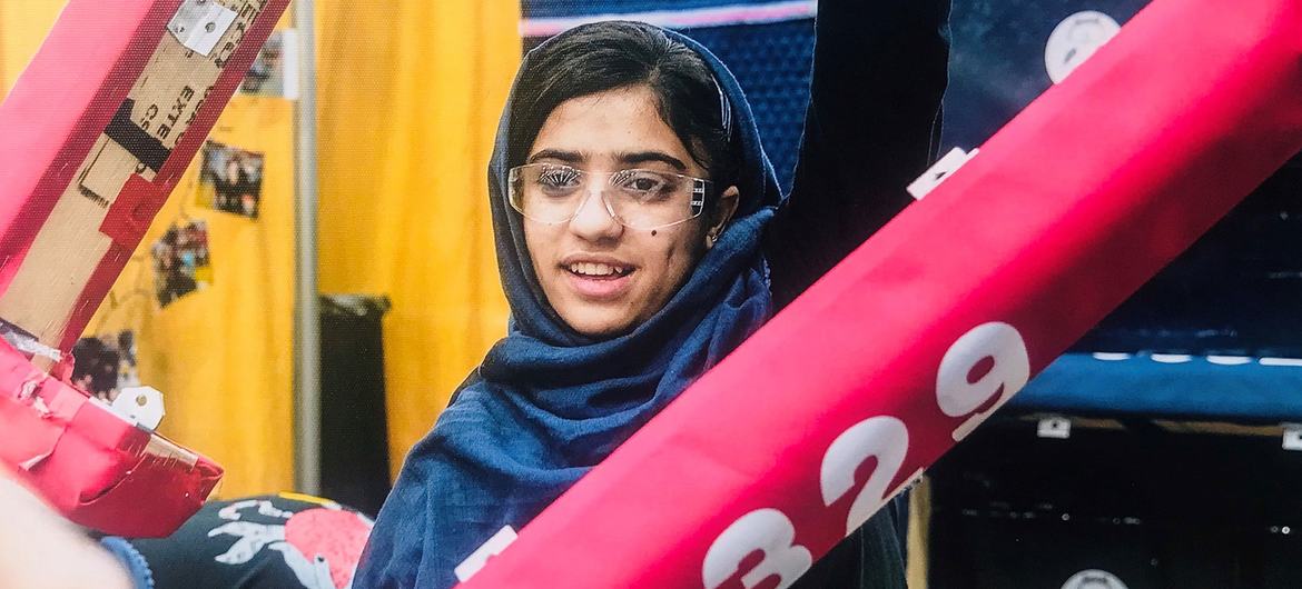 Somaya Faruqi, usando um hijab azul e óculos, sorri enquanto segura uma viga vermelha com o número 7329 em uma competição de robótica do FIRST Championship.
