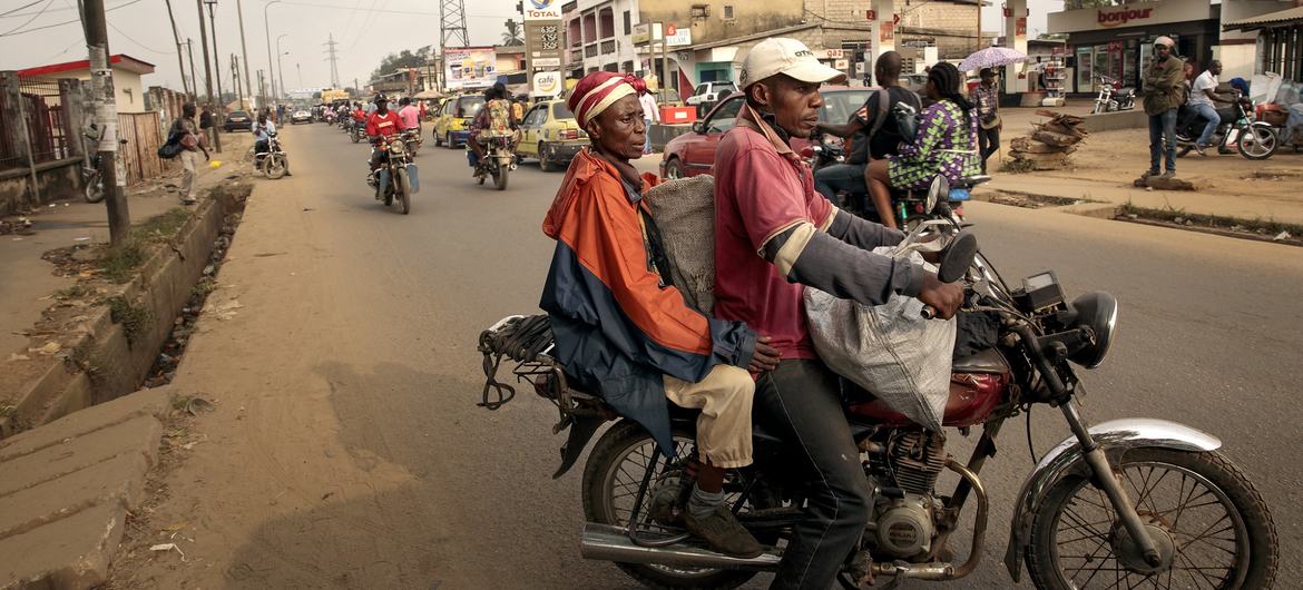 Dos hombres viajando en una motocicleta taxi por una calle de Camerún, con una gasolinera Total al fondo.