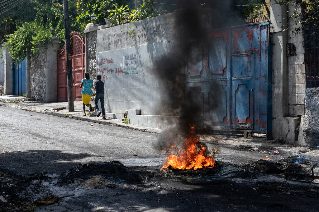 Moto unawaka kutoka matairi barabarani huko Port-au-Prince, Haiti, wakati wa maandamano. Watu wawili wanatembea kwenye njia ya watembea kwa miguu iliyo karibu.