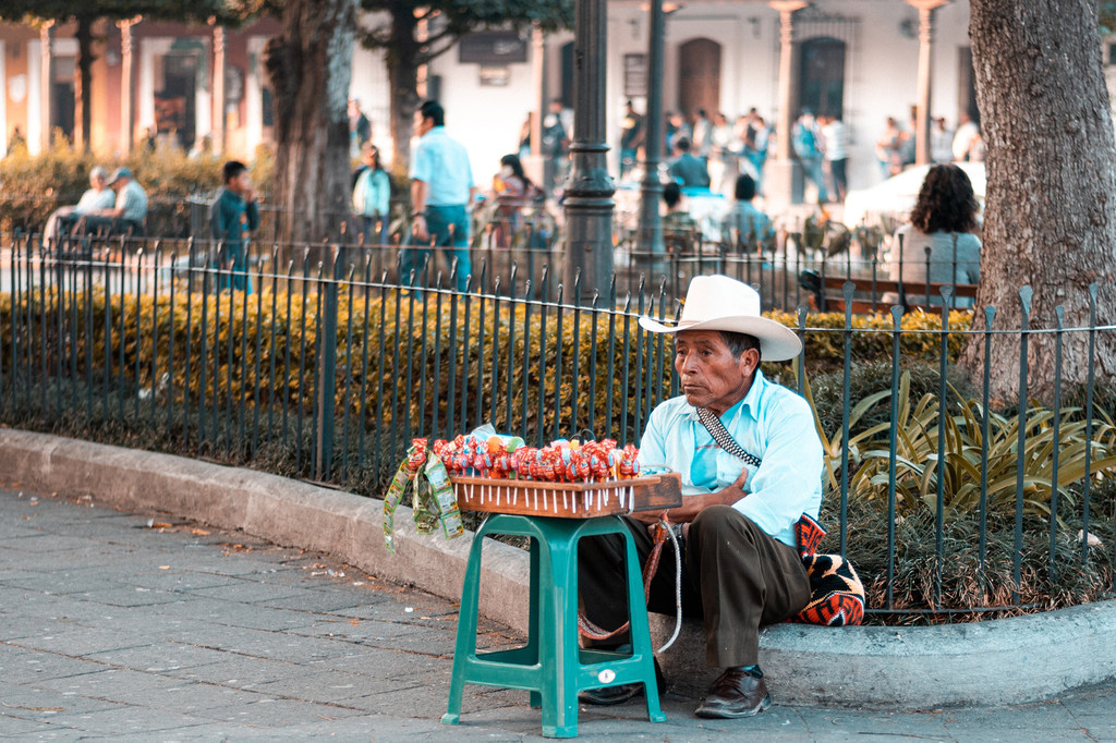 Un hombre con un sombrero blanco está sentado en la acera de una plaza en Antigua, Guatemala, con una bandeja de piruletas de colores sobre un taburete verde frente a él.