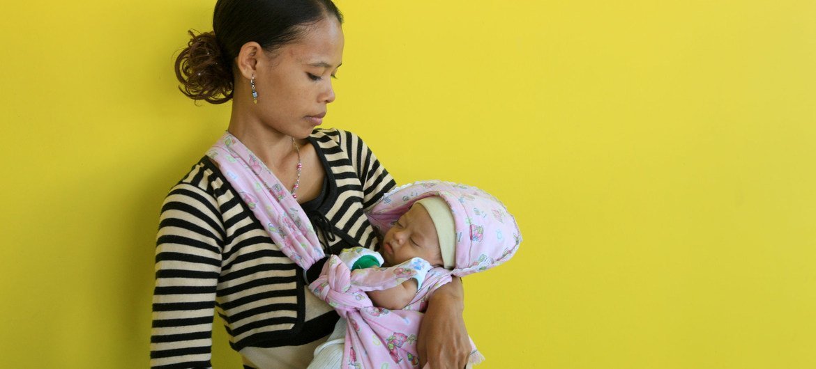 Uma mãe segura seu bebê recém-nascido em um sling rosa contra uma parede amarela.