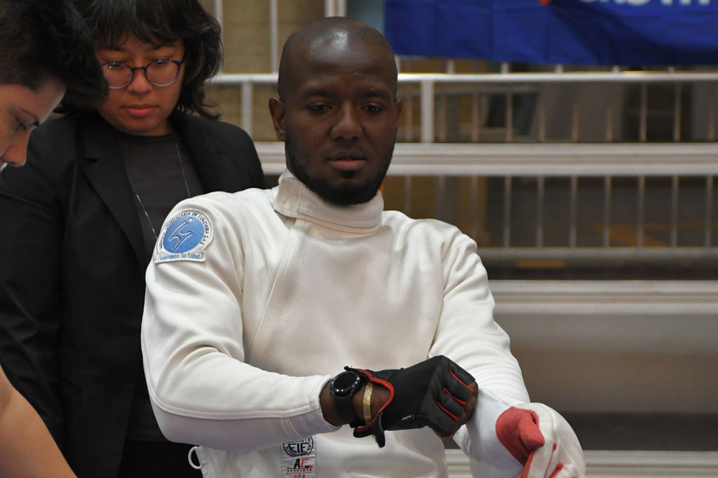 Un esgrimista, vestido con uniforme blanco y guantes negros, ajusta su equipo durante una competición. Otra persona, con chaqueta negra, observa la escena al fondo.