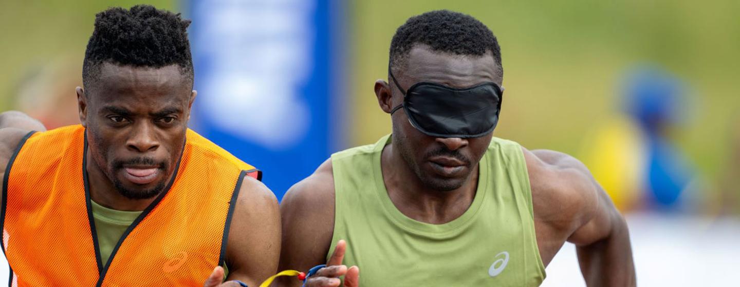 Un athlète handisport malvoyant, vêtu d'un débardeur vert et d'un masque noir pour les yeux, court avec son guide, qui porte un gilet de sécurité orange, pendant une course.