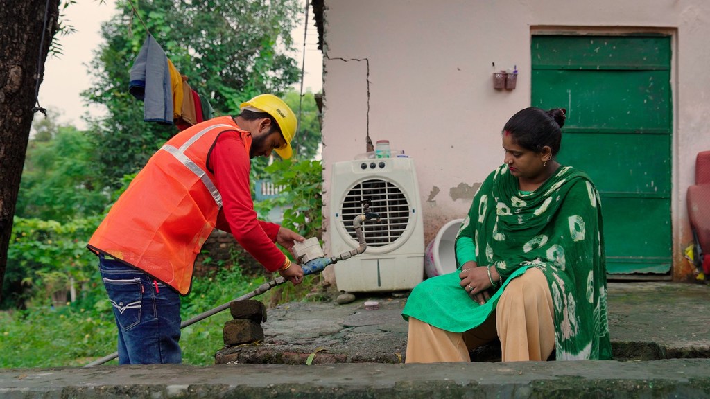भारत के उत्तराखंड राज्य के हरिद्वार में एक पुरुष तकनीशियन सुरक्षा जैकेट और हेलमेट पहनकर एक महिला के लिए पानी का पाइप ठीक कर रहा है।