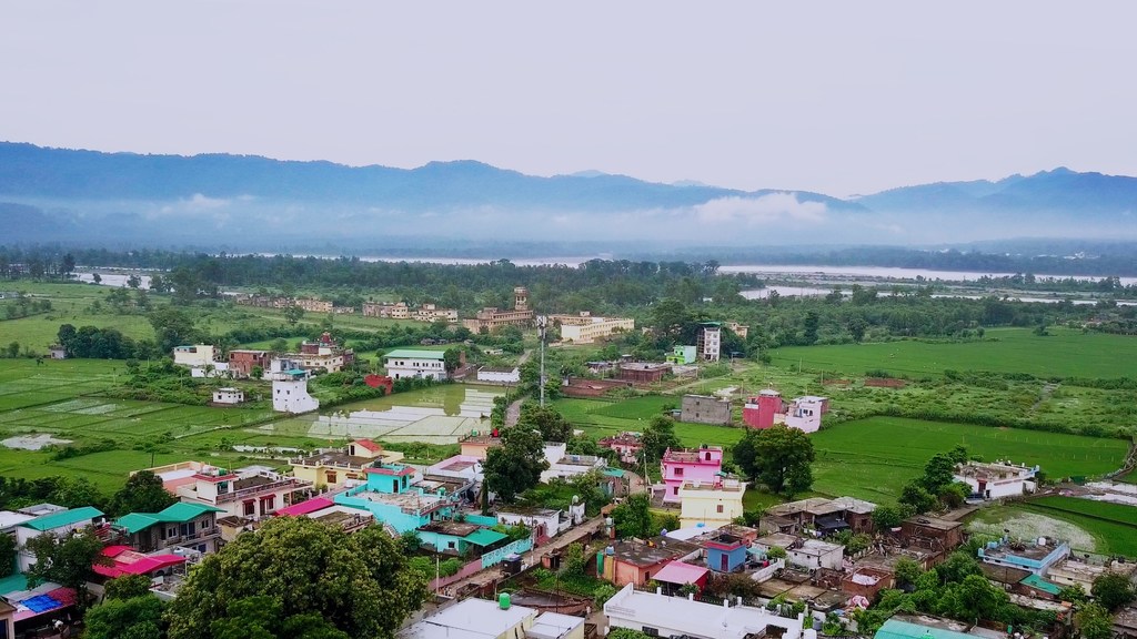भारत के उत्तराखंड में एक अर्ध-शहरी बस्ती का हवाई दृश्य, जिसमें पृष्ठभूमि में हरे-भरे कृषि क्षेत्र और पहाड़ दिखाई दे रहे हैं।