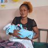 Uma mulher de cabelos cacheados está sentada em uma cama de hospital segurando um bebê recém-nascido enrolado em uma manta azul.