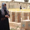 امرأة ترتدي حجاباً أزرق تقف في منطقة توزيع المواد الغذائية في دير حافر بسوريا، بينما يقوم موظفو WFP الأحمر السوري، وهم يرتدون الكمامات، بتحضير صناديق المساعدات.