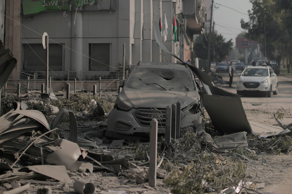 Un coche dañado yace rodeado de escombros en una calle de Gaza, con banderas palestinas ondeando en un edificio al fondo.