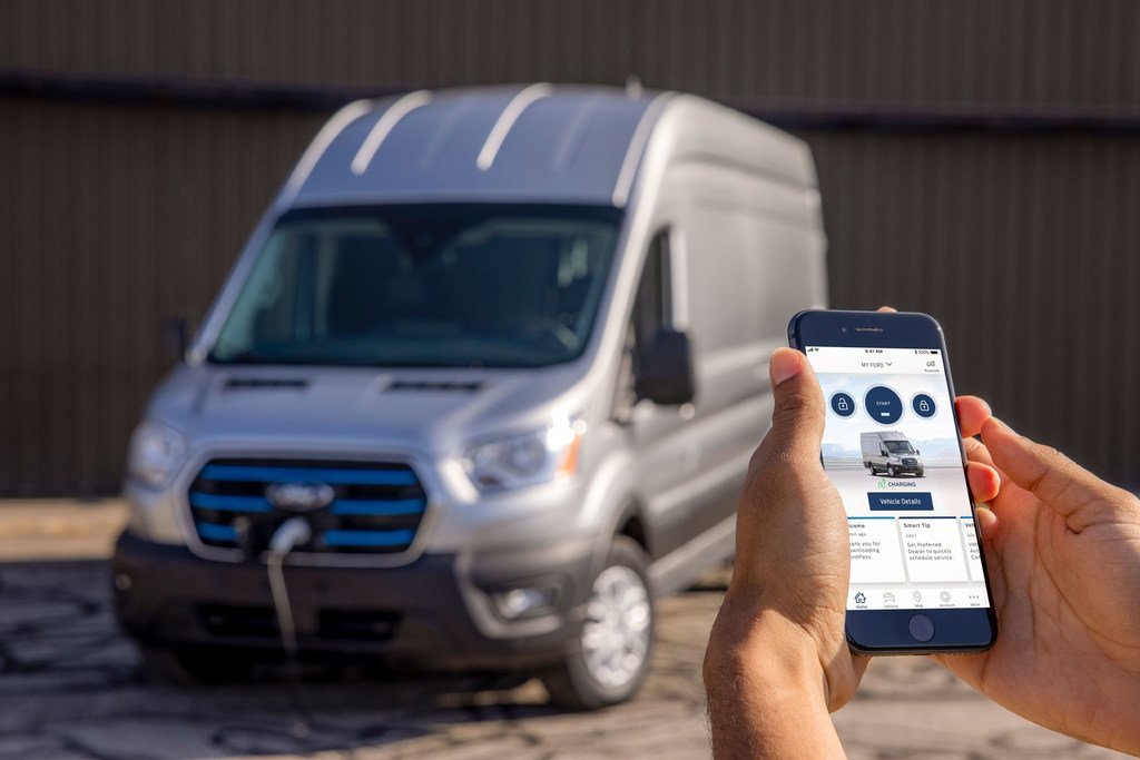 Una furgoneta de carga eléctrica Ford E-Transit plateada está conectada a una estación de carga, mientras una persona sostiene un teléfono inteligente que muestra los detalles del vehículo en una aplicación móvil.