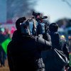 Um cinegrafista de jaqueta escura segura uma grande câmera de vídeo sobre o ombro, filmando um evento ao ar livre em um dia ensolarado.