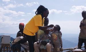 Mfanyakazi wa afya aliyevaa shati la njano akimpa mtoto mdogo chanjo ya polio huko West Pokot, Kenya, huku mandhari ya milima ikiwa nyuma yake.