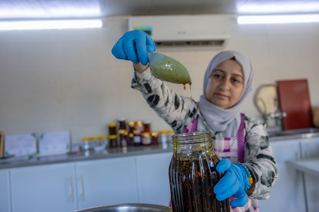 A Syrian refugee woman in a kitchen fills a glass jar with jam while wearing blue gloves and an apron.