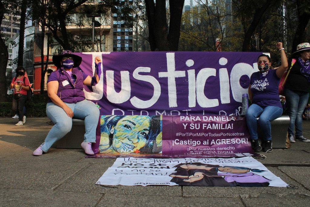 Mujeres mexicanas con máscaras y camisas moradas se sientan en un banco sosteniendo una gran pancarta morada en la que se lee «Justicia» (Justicia) en una protesta contra los feminicidios. En la pancarta se muestra un retrato pintado de una mujer llamada Daniela.