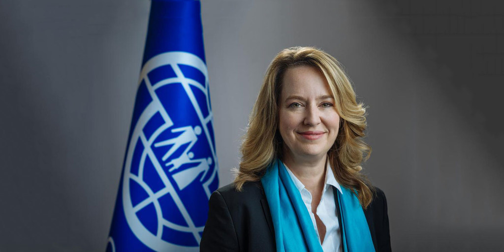 Retrato de Amy Pope, la primera mujer Directora General de la Organización Internacional para las Migraciones (OIM), de pie frente a la bandera de la OIM.