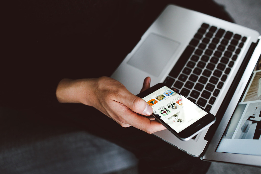 La mano de una persona sostiene un teléfono inteligente que muestra una aplicación de redes sociales junto a una computadora portátil abierta.
