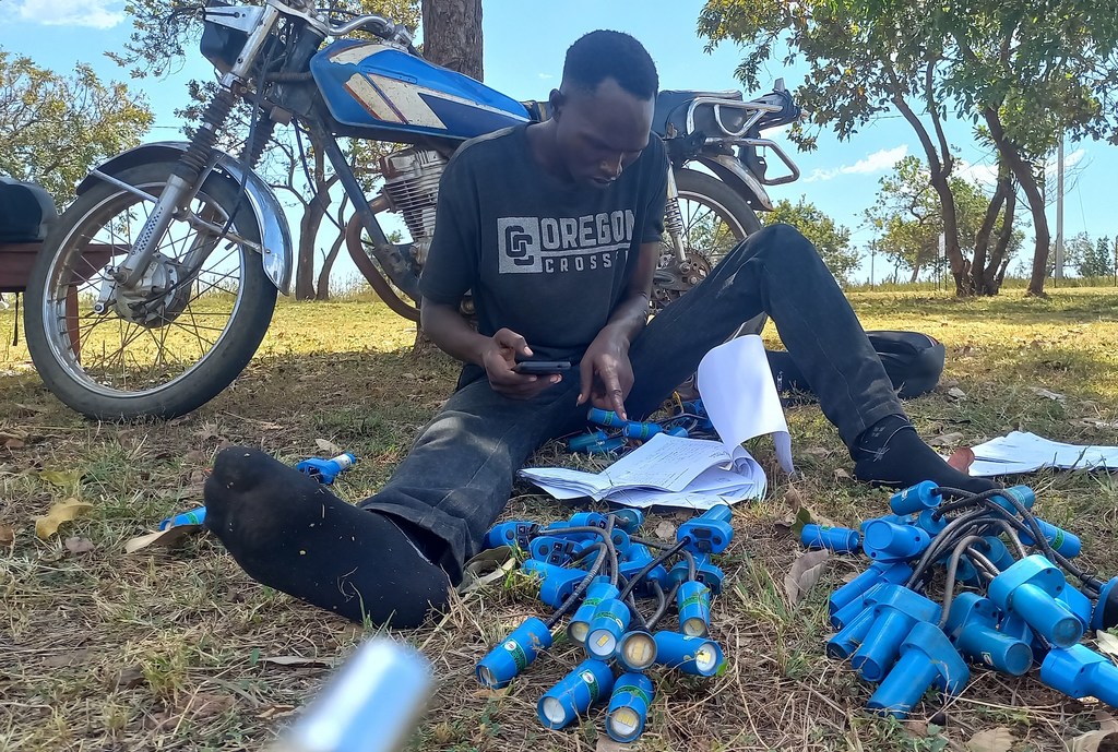 Mhamasishaji wa jamii katika Makazi ya Wakimbizi ya Bidibidi nchini Uganda anapitia taa za jua zilizovunjika kwa ajili ya matengenezo, huku pikipiki ikiwa imeegeshwa karibu.