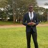 Mwanamume aliyevaa suti na miwani ya jua amesimama kwenye nyasi yenye nyasi huku miti na bendera zikiwa nyuma.