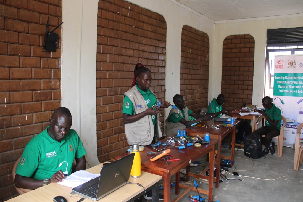 Mafundi waliovaa sare za kijani za IOM wanafanya kazi katika meza za kutengeneza vifaa vya kielektroniki katika Kituo cha Ubunifu cha Mercy Corps huko Bidibidi, Uganda.