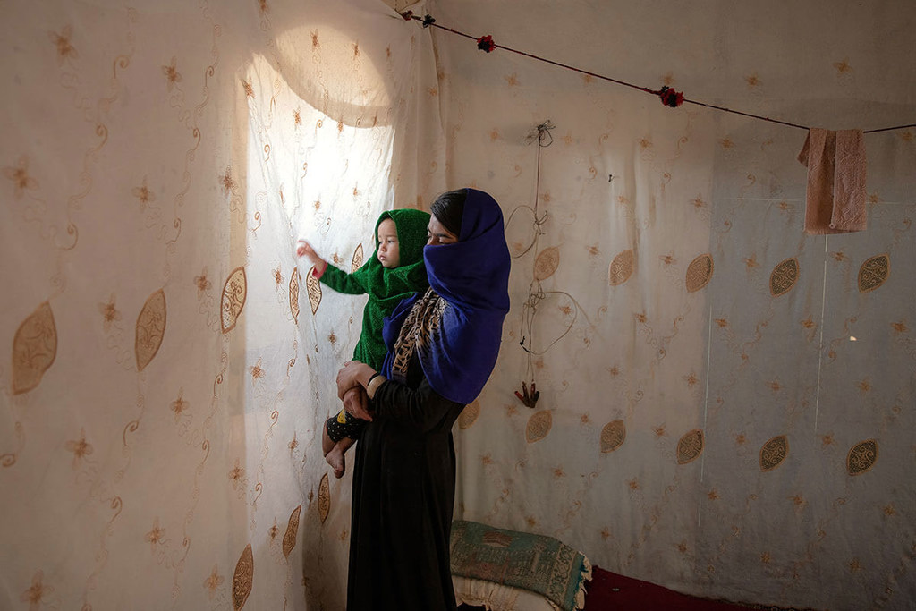 Une mère afghane tient sa fille dans ses bras, fixant la lumière derrière sa fenêtre obscurcie.