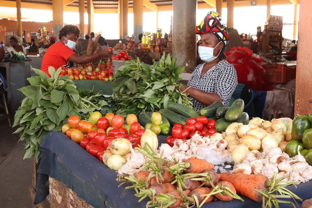 Duas mulheres usando máscaras vendem produtos frescos em uma barraca de mercado em Luanda, Angola. A barraca está repleta de tomates, cenouras, pepinos, cebolas, alho e verduras.