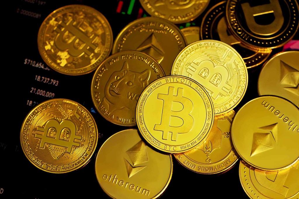 Una colección de monedas físicas de color oro, incluyendo Bitcoin, Ethereum y Dogecoin, mostradas en una pantalla que muestra datos financieros.