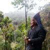 Um homem indígena com um cocar de penas está em uma floresta tropical, segurando uma pequena muda de árvore.