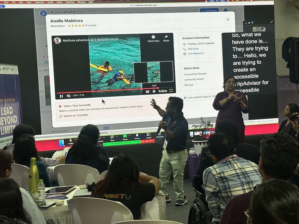 एक व्यक्ति सम्मेलन में मंच पर प्रस्तुति दे रहा है, उसके बगल में एक सांकेतिक भाषा का दुभाषिया है और उसके सामने दर्शक बैठे हैं।