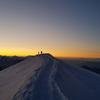 Silhuetas de dois caminhantes em pé sobre uma cordilheira nevada ao nascer do sol, com o Mont Blanc ao fundo.
