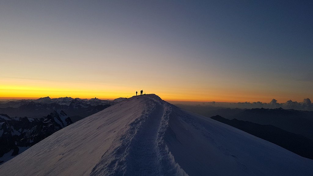 Silhuetas de dois caminhantes em pé sobre uma cordilheira nevada ao nascer do sol, com o Mont Blanc ao fundo.