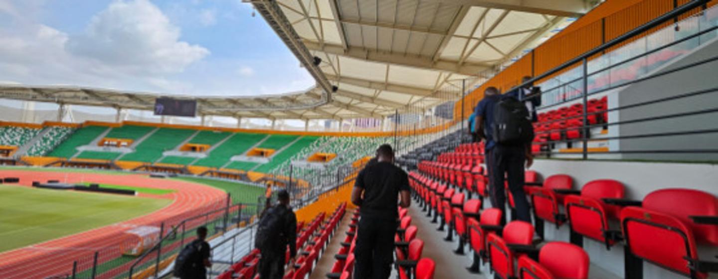 Personal de seguridad camina junto a una fila de asientos rojos en un estadio vacío, con una pista de atletismo y un campo verde al fondo.