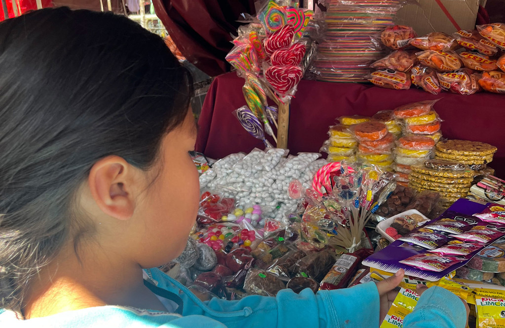 Una niña con una camisa azul observa una colorida exhibición de caramelos y dulces en un puesto del mercado.