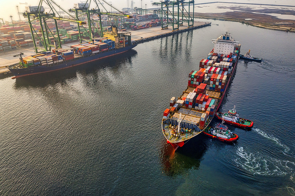 Vista aérea de dois navios porta-contentores em um porto movimentado, um deles sendo guiado por rebocadores.