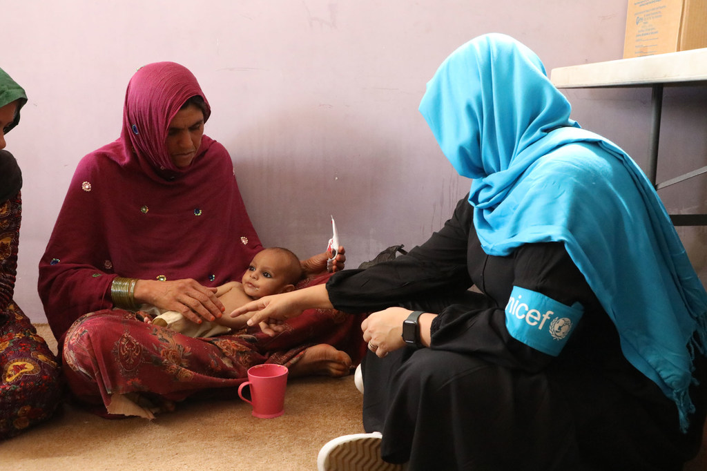 Mfanyakazi UNICEF aliyevaa kitambaa cha bluu kichwani na gauni jeusi, akiwa na kitambaa cha mkono UNICEF , anachunguza mkono wa mtoto huku bibi yake mtoto akimtazama. Kikombe cha waridi kimewekwa sakafuni karibu.