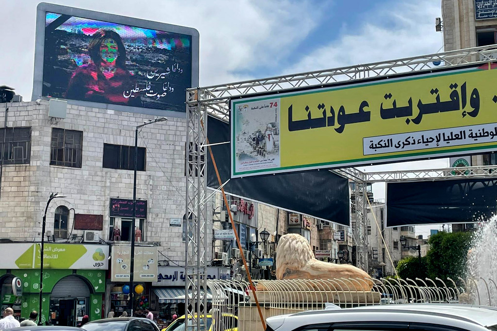مشهد من ساحة المنارة في رام الله، يضم تمثالاً لأسد، ونافورة، ولوحة إعلانية رقمية كبيرة، ولافتة إعلانية مكتوب عليها نص عربي.