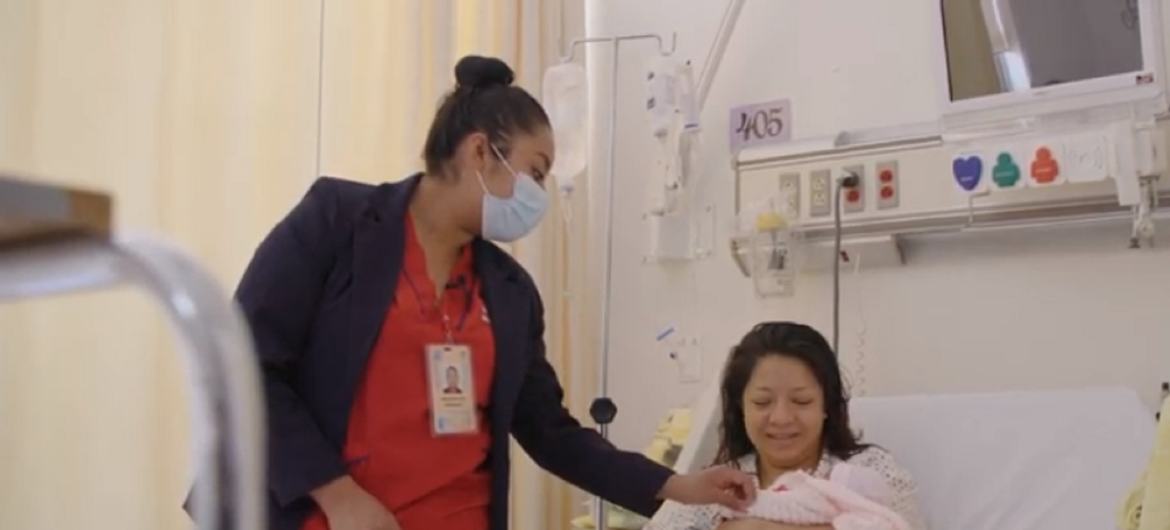 Una matrona permanece de pie junto a una madre en una cama de hospital, que sostiene a su bebé recién nacido.