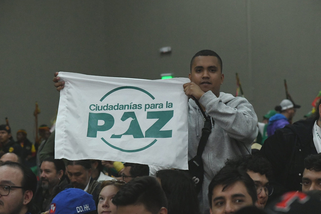 Un hombre sostiene una bandera blanca con texto verde que dice 'Ciudadanías para la PAZ' en un evento en un espacio cerrado con una multitud de personas.
