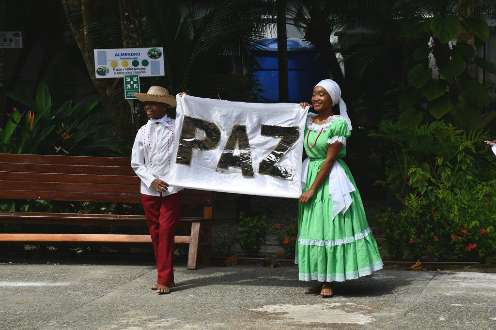 Two young people, a male and a female, hold a white banner with the word PAZ written on it. The male is wearing a traditional hat, a white shirt, and red pants. The female is wearing a traditional green dress and a white headscarf. They are standing outdoors on a paved area with a bench and plants in the background.