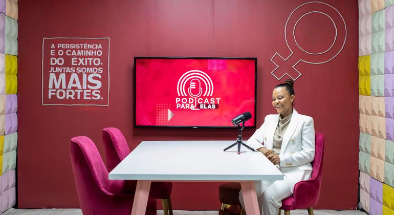 A ativista social Alima está sentada em um estúdio de podcast, sorrindo enquanto está sentada à mesa com um microfone. Uma tela atrás dela exibe o logotipo do 'Podcast Para Elas'.