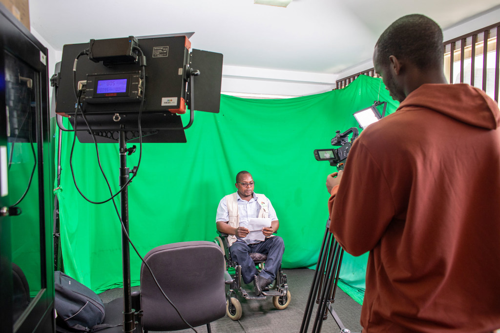 O jornalista Abdul Remane Ibraimo, em uma cadeira de rodas, segurando papéis, em um cenário com tela verde, sendo filmado por um cinegrafista.