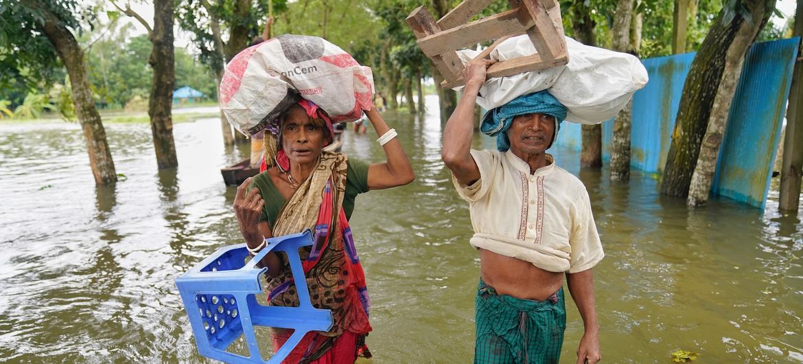 बांग्लादेश के फेनी में एक पुरुष और एक महिला बाढ़ के पानी में से गुजरते हुए अपने सिर पर प्लास्टिक की कुर्सी, लकड़ी का स्टूल और सामान से भरी बोरियां जैसी बचाई गई चीजें ले जा रहे हैं।